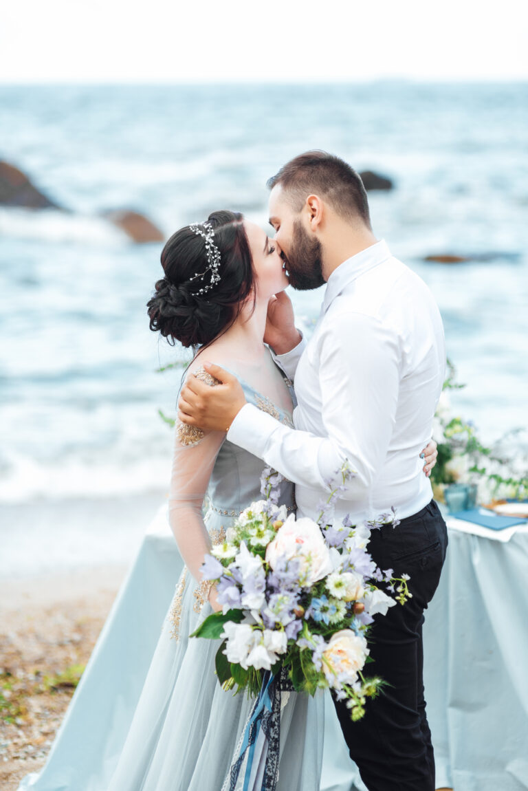 couple with a bride in a blue dress walk along the ocean shore